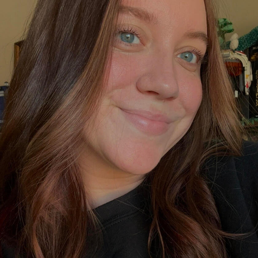 This image shows a woman with light skin and shoulder-length, wavy brown hair. She is wearing a black top and has a soft smile, with blue-green eyes looking slightly to the side. In the background, there is a well-lit room with a bookshelf containing books